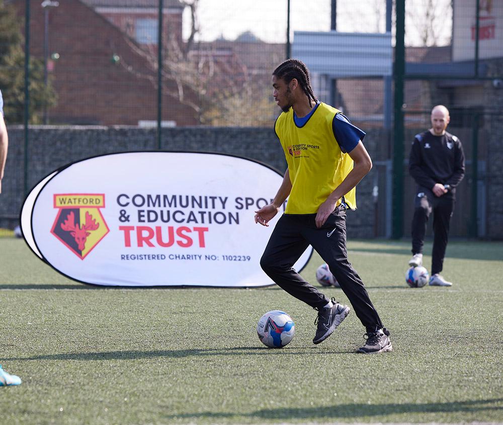 Man controls football on 4G pitch while coach watches on, banner with Watford FC crest reading Community Sport and Education Trust