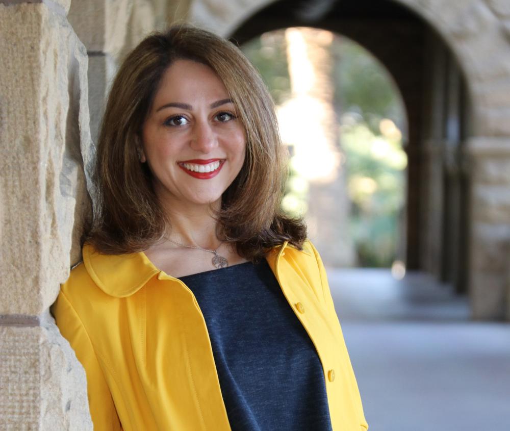 A portrait of Dr. Sara Nasserzadeh in a yellow coat in front of an old building with an archway