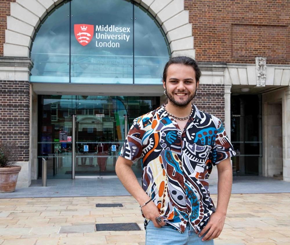 Omar Maaouan Veiga in a colourful shirt, standing and smiling outside the entrance to Middlesex University's College Building