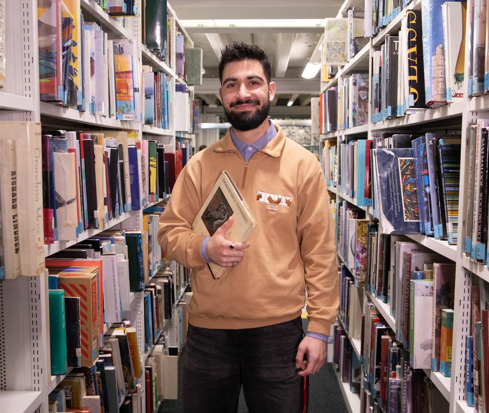 A man carrying a textbook whilst standing between bookshelves