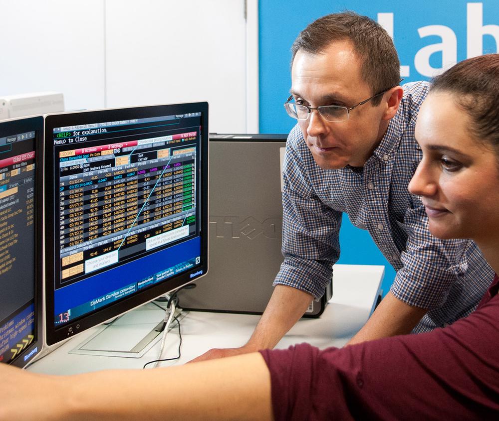 A student sits at a desk looking at financial data as a lecturer looks on