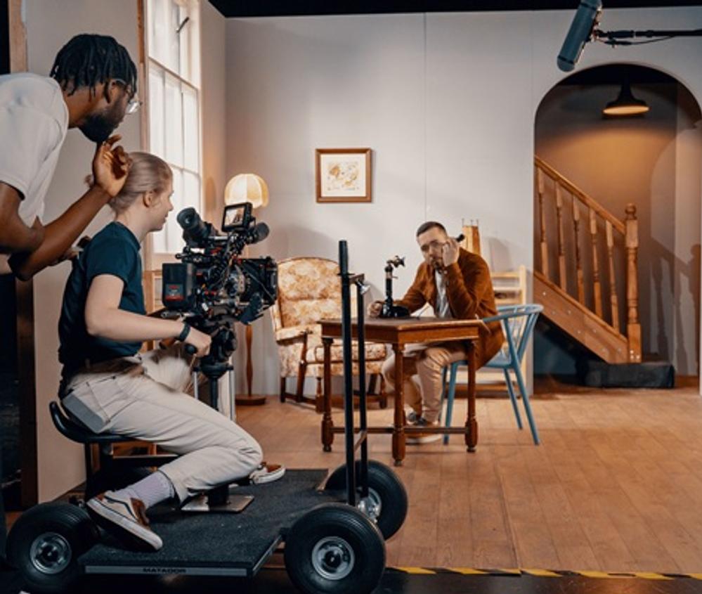 Students using a film camera to film an actor on set who is sitting at a table using a phone
