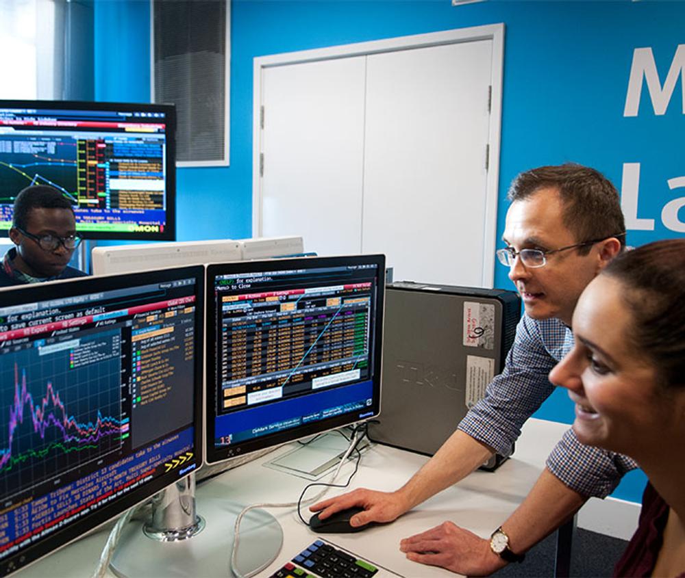 a student and professor sitting at a desk with screens review data in the financial markets lab