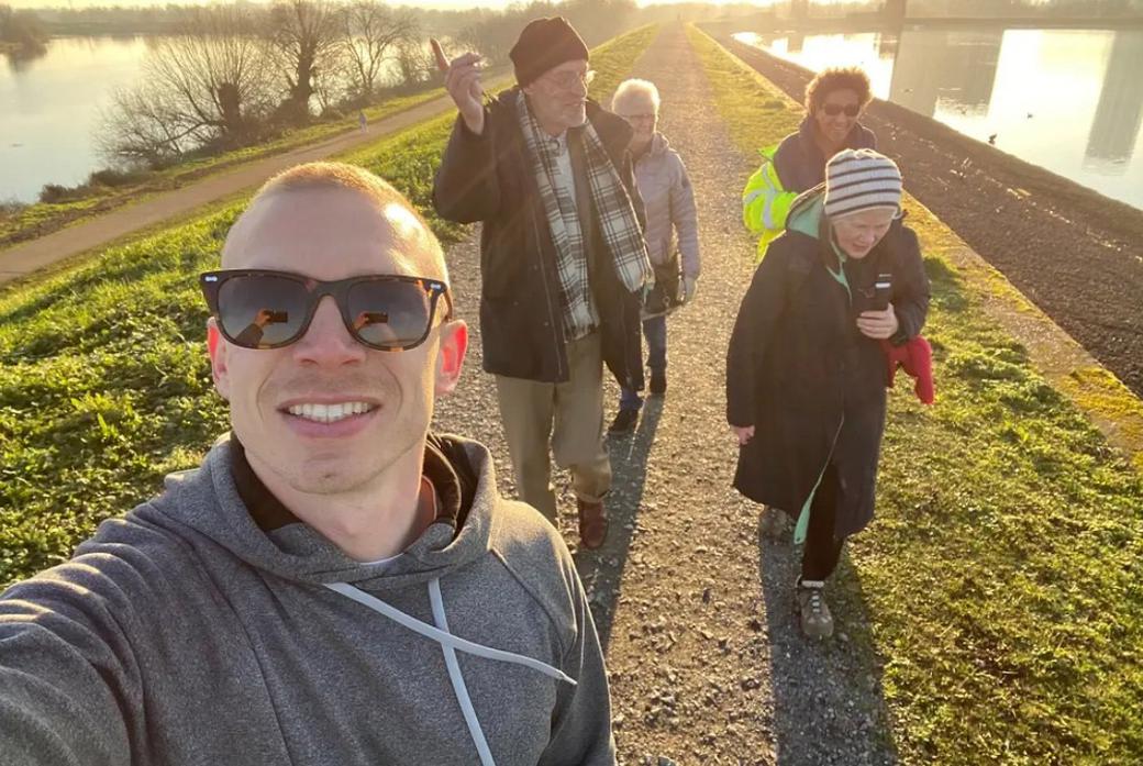 A young man in a hoodie and sunglasses takes a selfie in front of a group of older people in hats and scarves. They seem to be in the middle of a walk