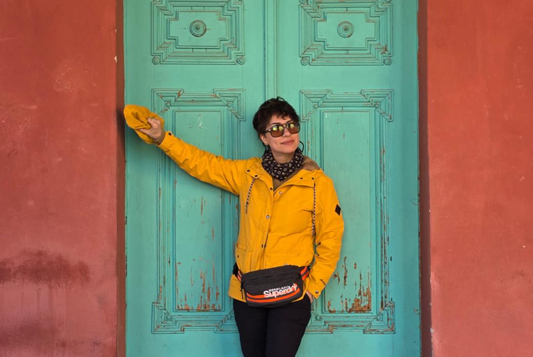 A woman wearing a yellow jacket and sunglasses poses in front of a jade green door