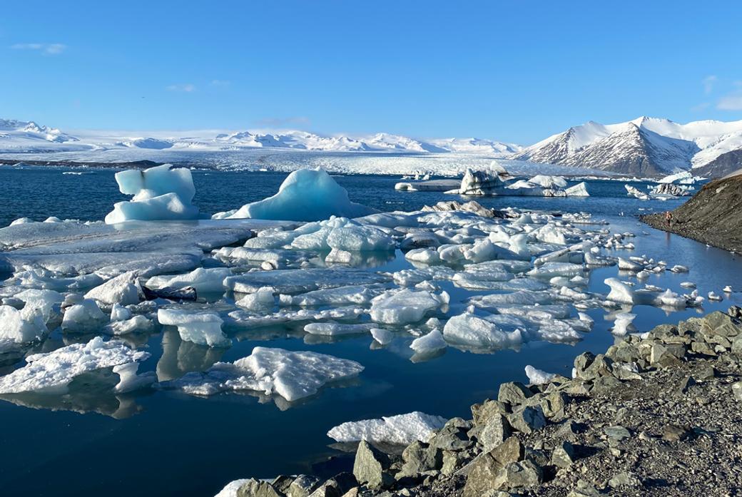 A view of the arctic ocean with rocks in the foreground, a blue sky and ice floes in the sea