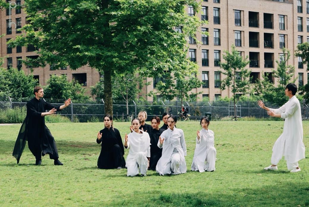 A group of dancers in black and white costumes flowing costumes strike dramatic poses in Colindale Park