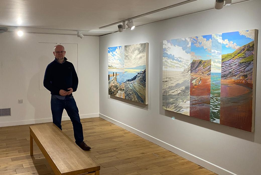 A portrait of Dr John Timberlake standing next to his paintings in the Granary Gallery, Berwick-Upon-Tweed