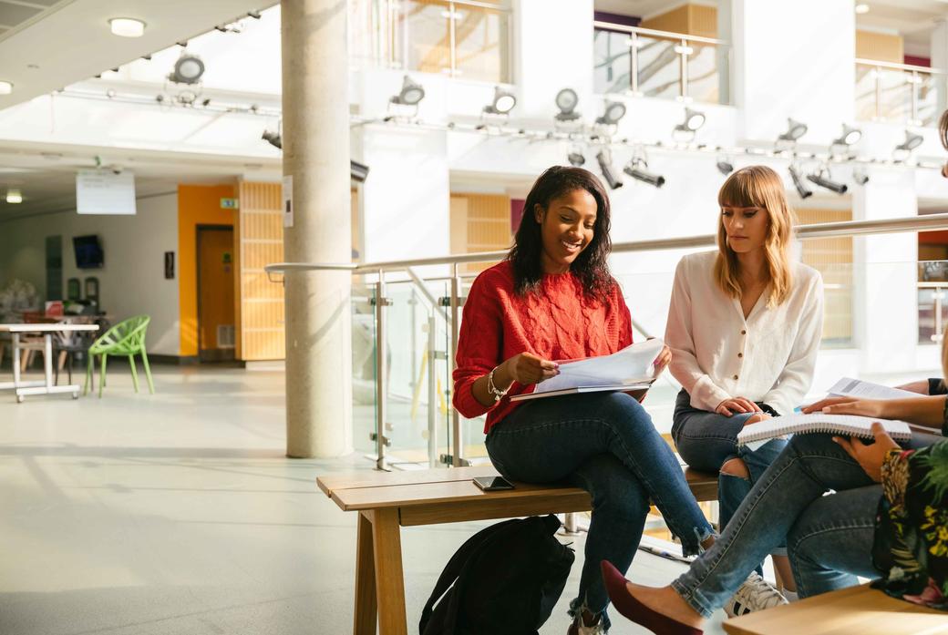 Three students sitting on a bench inside a building, engaged in conversation and studying together.