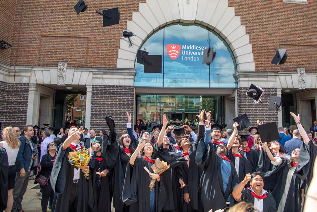 A group of graduates in gowns throw their mortar boards into the air to celebrate graduation in front of a 91���� building
