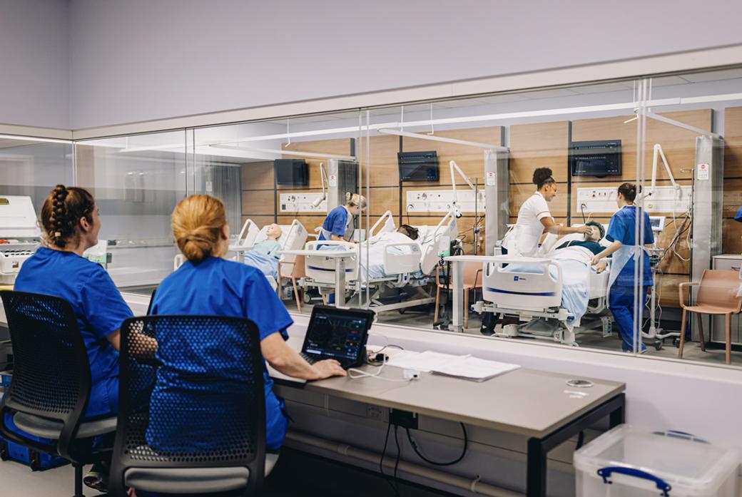 a nursing station in a hospital ward with two seated nurses observing the care of patients