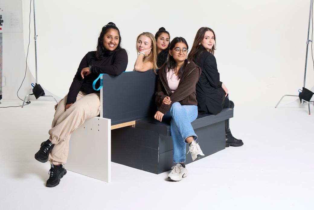A group of five women sitting on a bench against a white photographic studio backdrop