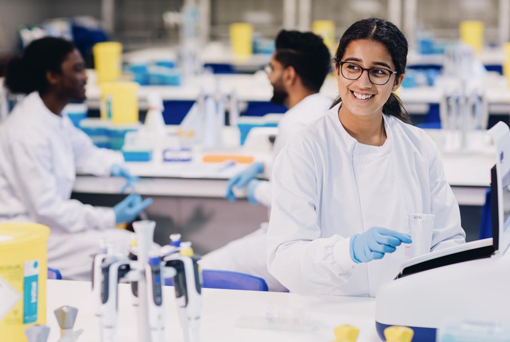 A smiling woman works in a science lab with two people chatting behind her