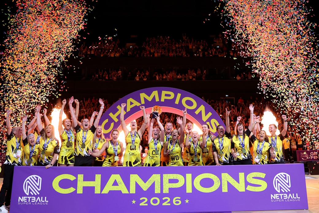 A netball team celebrates behind a sign saying 'champions' and a firework explosion a each side