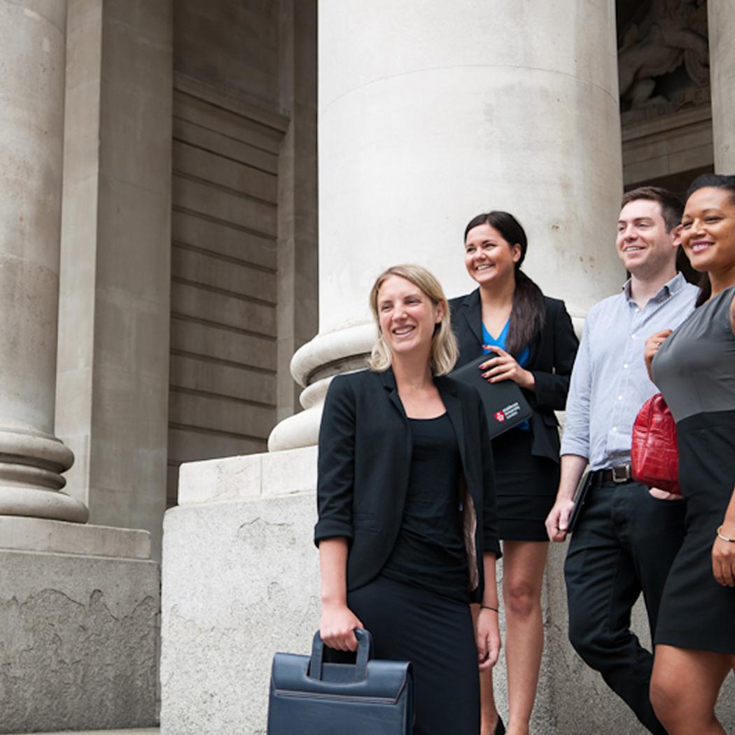 Four professionally dressed students stand outside a building with large stone columns, smiling and holding folders and bags.