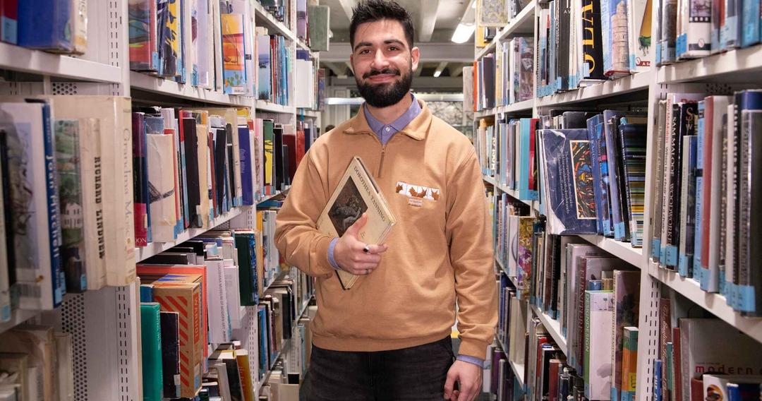 A man carrying a textbook whilst standing between bookshelves