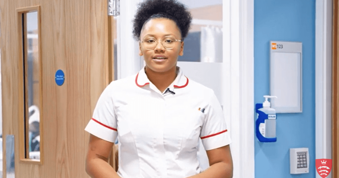 A student nurse in uniform stands in a hospital corridor, talking to the camera