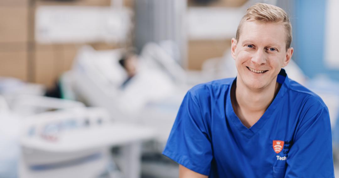 A male nurse in blue scrubs sitting at a desk, reviewing patient charts.