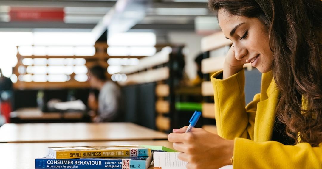A woman seated at a table, engrossed in her notebook and pen, engaged in Business and Marketing studies.