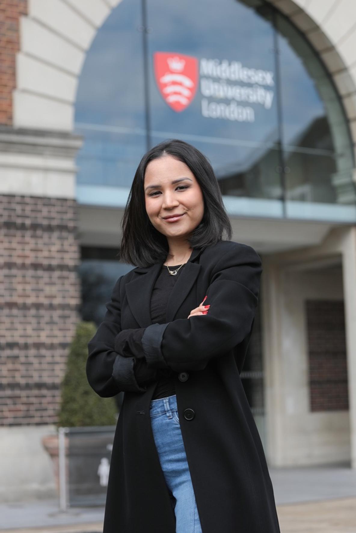 A smiling woman folding her arms, standing confidently in front of the entrance to Middlesex University London's Quad building