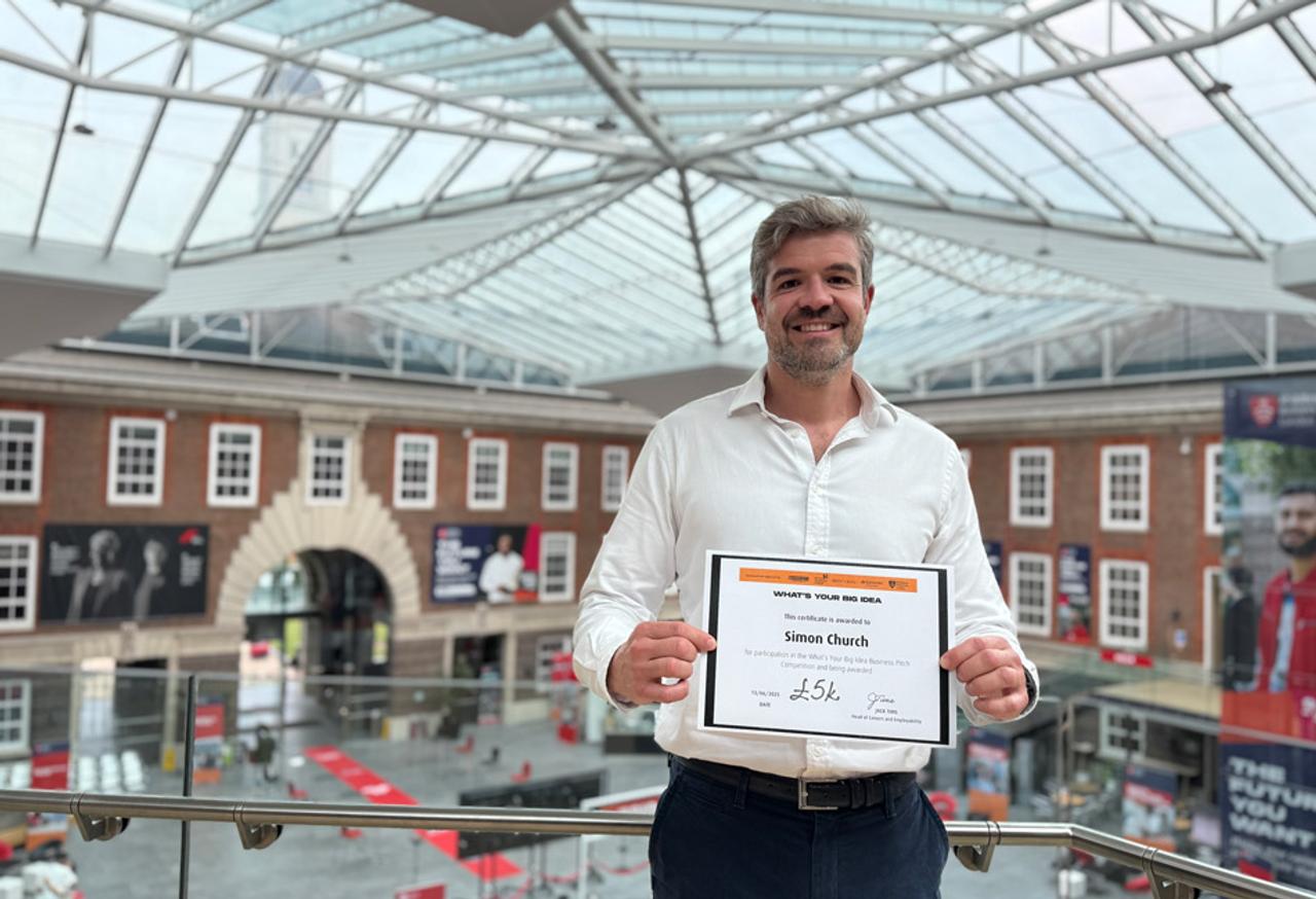 A MDX MBA student in a white shirt stands holding a certificate on the second floor balcony of the Quad