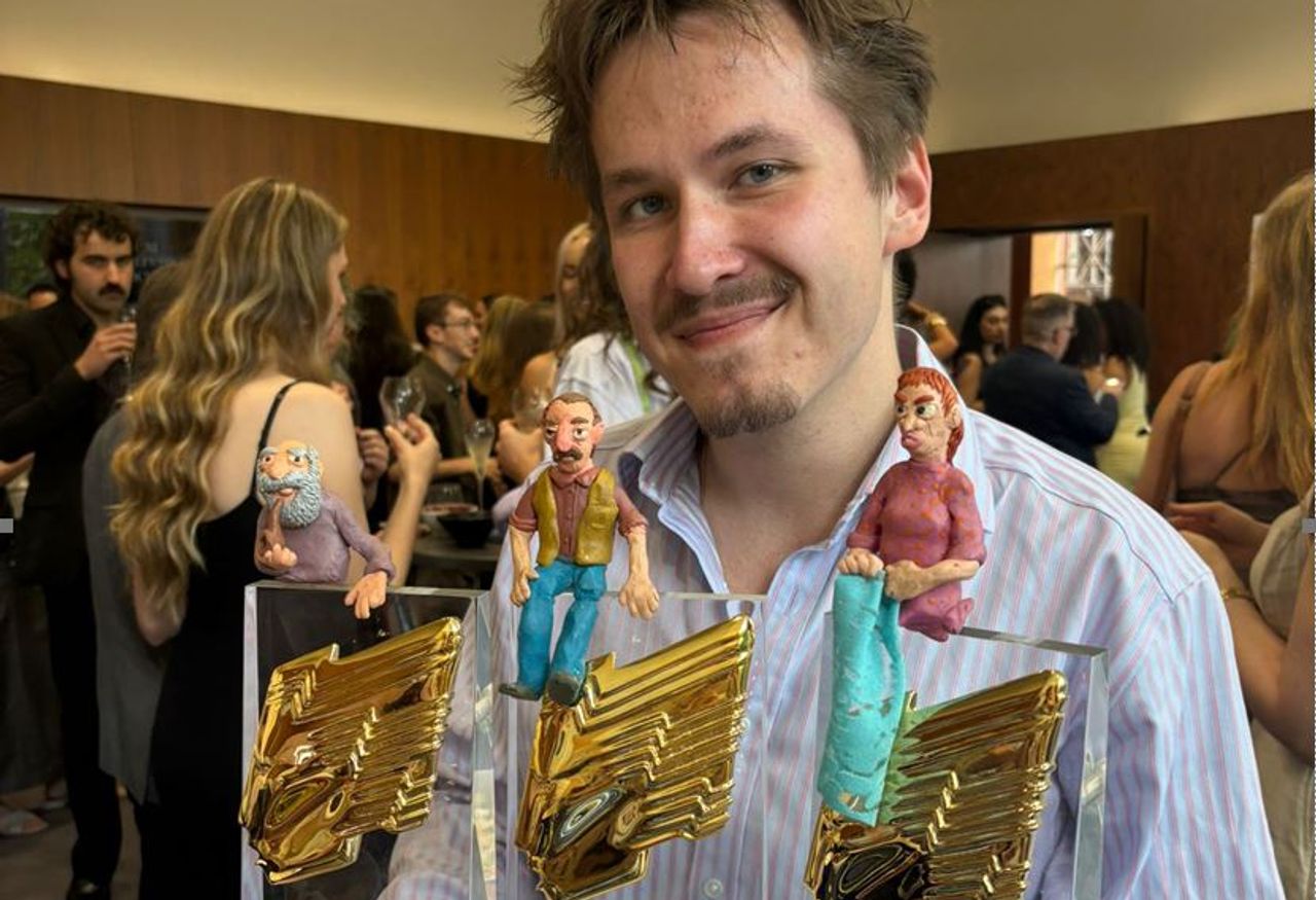 A Middlesex graduate with a light beard and moustache smiles in front of three awards. Plasticine model figures from his animation sit on top of the awards