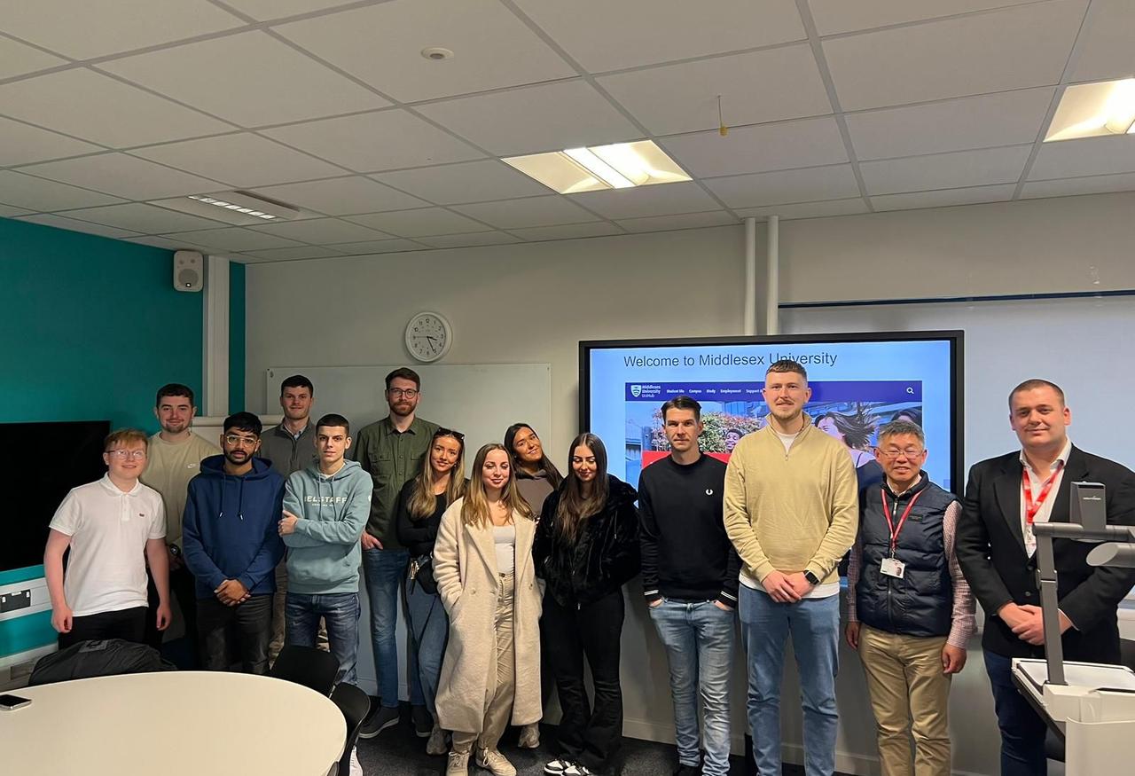 A group of young male and female students standing in a line, wearing casual clothes, in front of a large screen