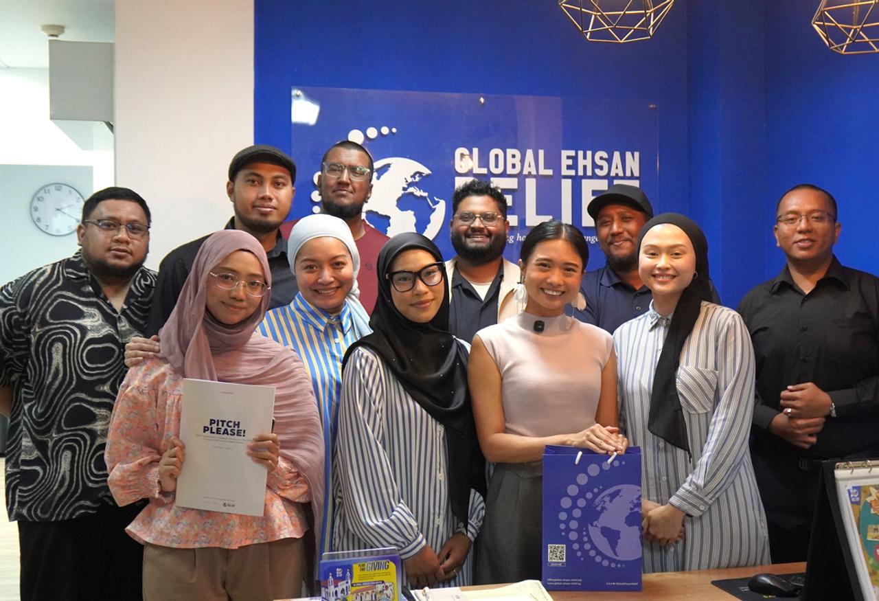 A group of men and women at Muslim anti-poverty charity Global Ehsan Relief stand in a group against a blue wall with their charity logo on it, smiling at the gamera