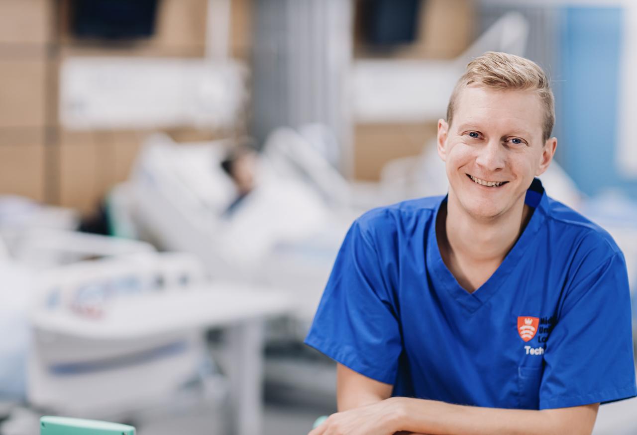 A male nurse in blue scrubs sitting at a desk, reviewing patient charts.