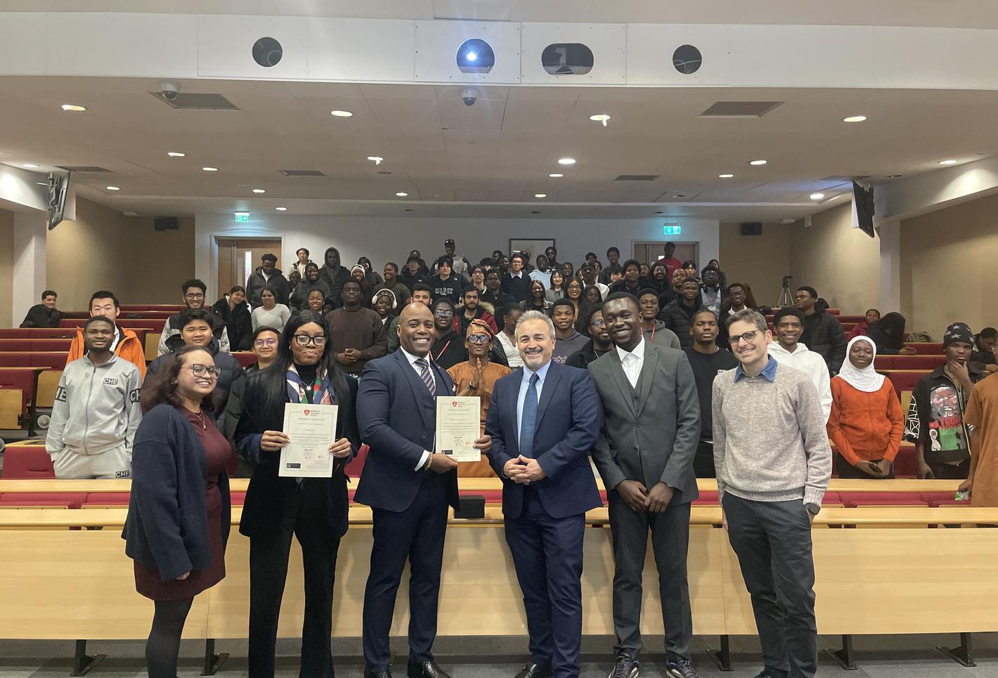 A line-up of Computer Science lecturers and staff stand with two Nigerian visitors to Middlesex in a lecture theatre in Hatchcroft building