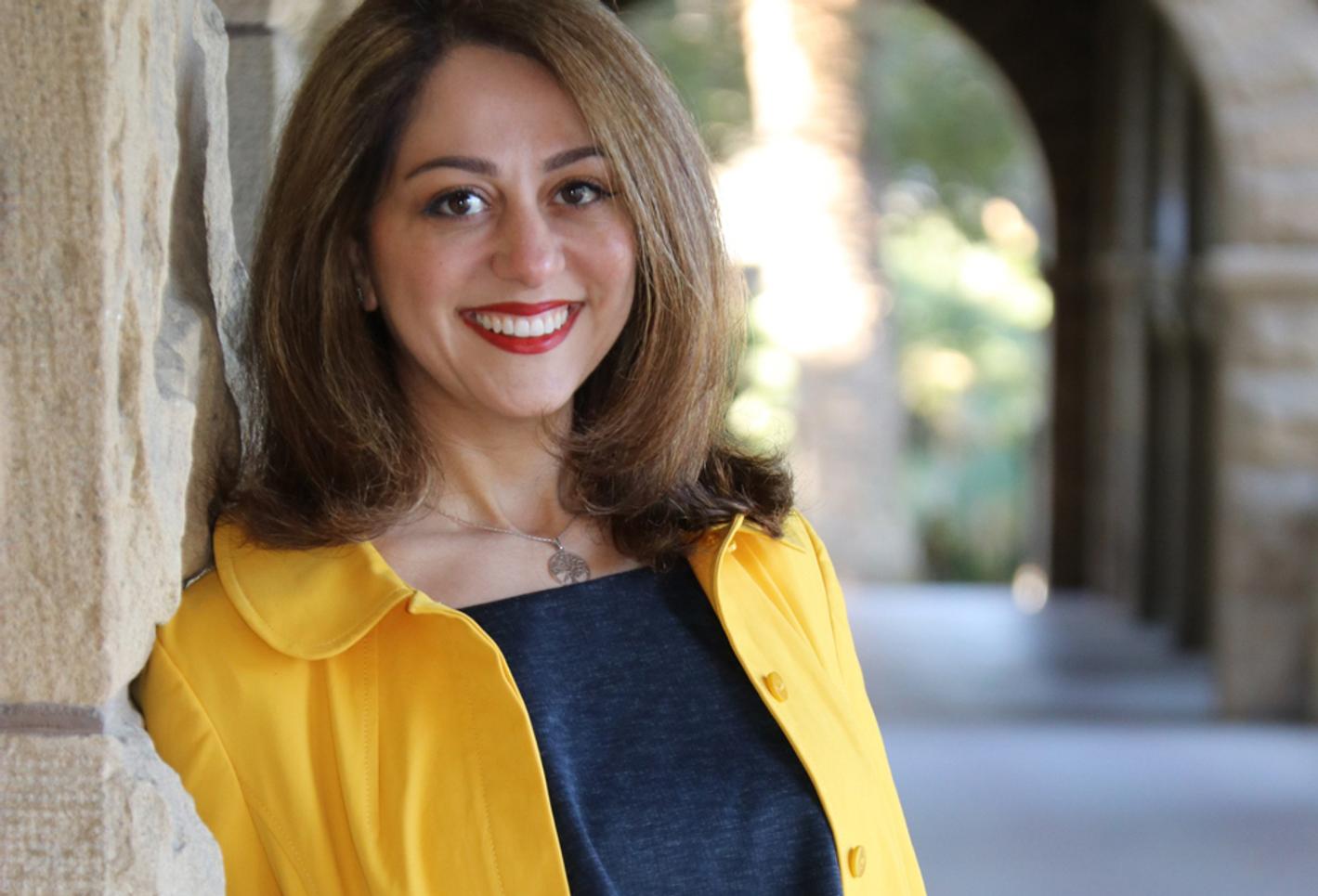 A portrait of Dr. Sara Nasserzadeh in a yellow coat in front of an old building with an archway