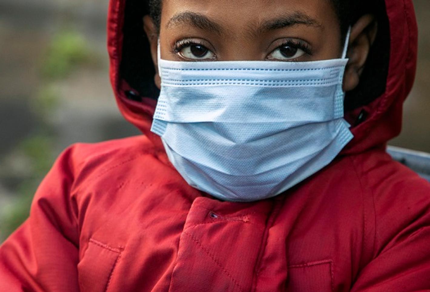 A photograph which is part of an art exhibition of a young black person with a blue surgical facemask on, wearing a red anorak with a hood, staring at the viewer