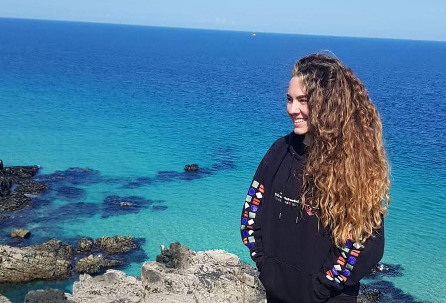 A young smiling woman dressed in black stands on a cliff next to the sea on a sunny day
