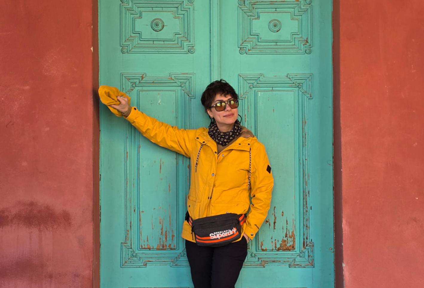 A woman wearing a yellow jacket and sunglasses poses in front of a jade green door