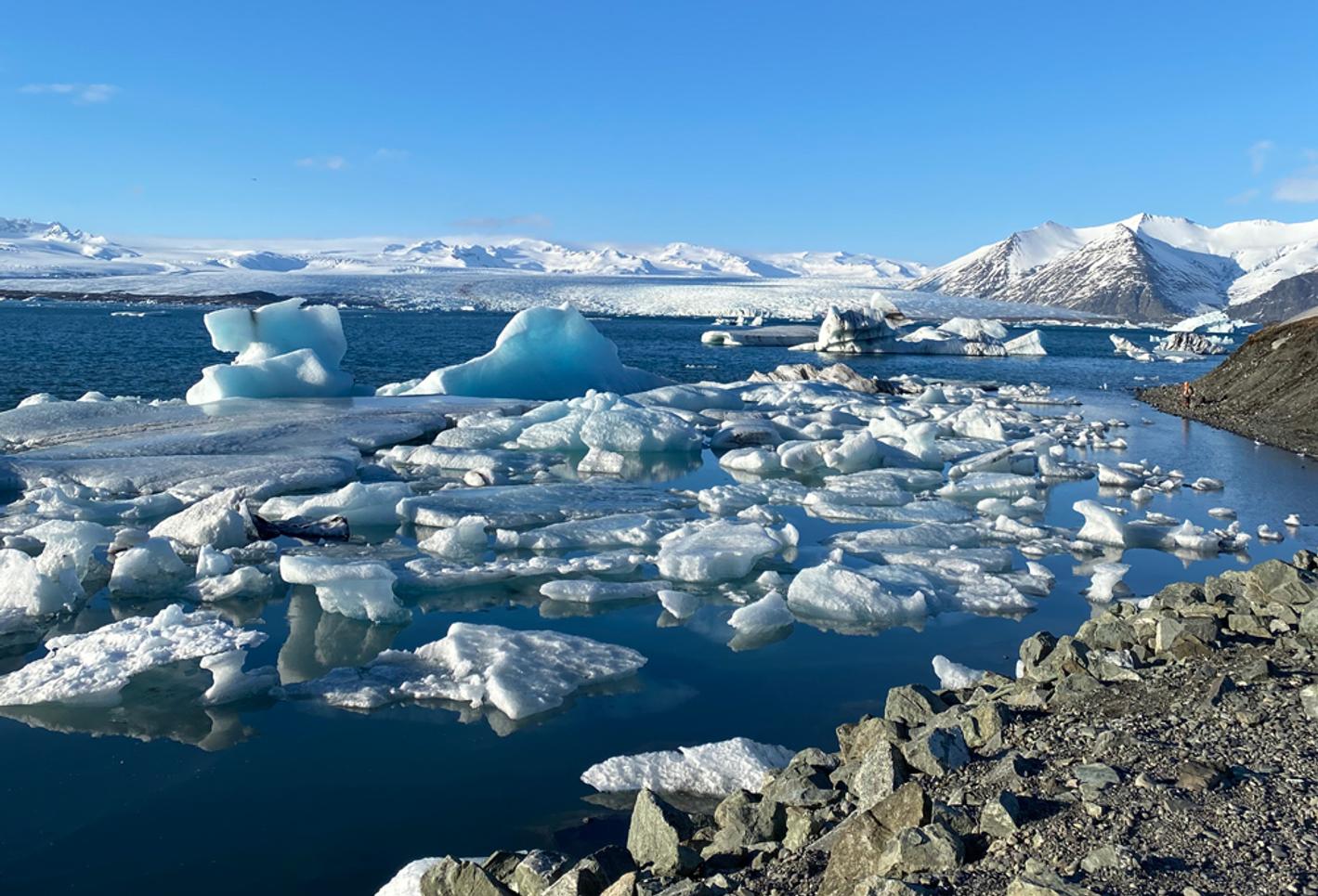 A view of the arctic ocean with rocks in the foreground, a blue sky and ice floes in the sea
