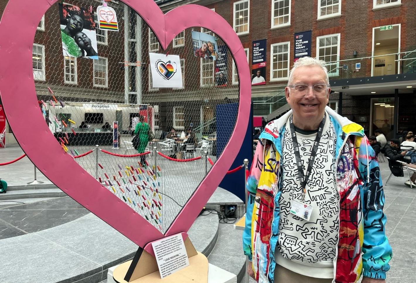 An older white man with white hair in a bright coloured graffiti-style jacket stands by a large cut-out sculpture of a heart