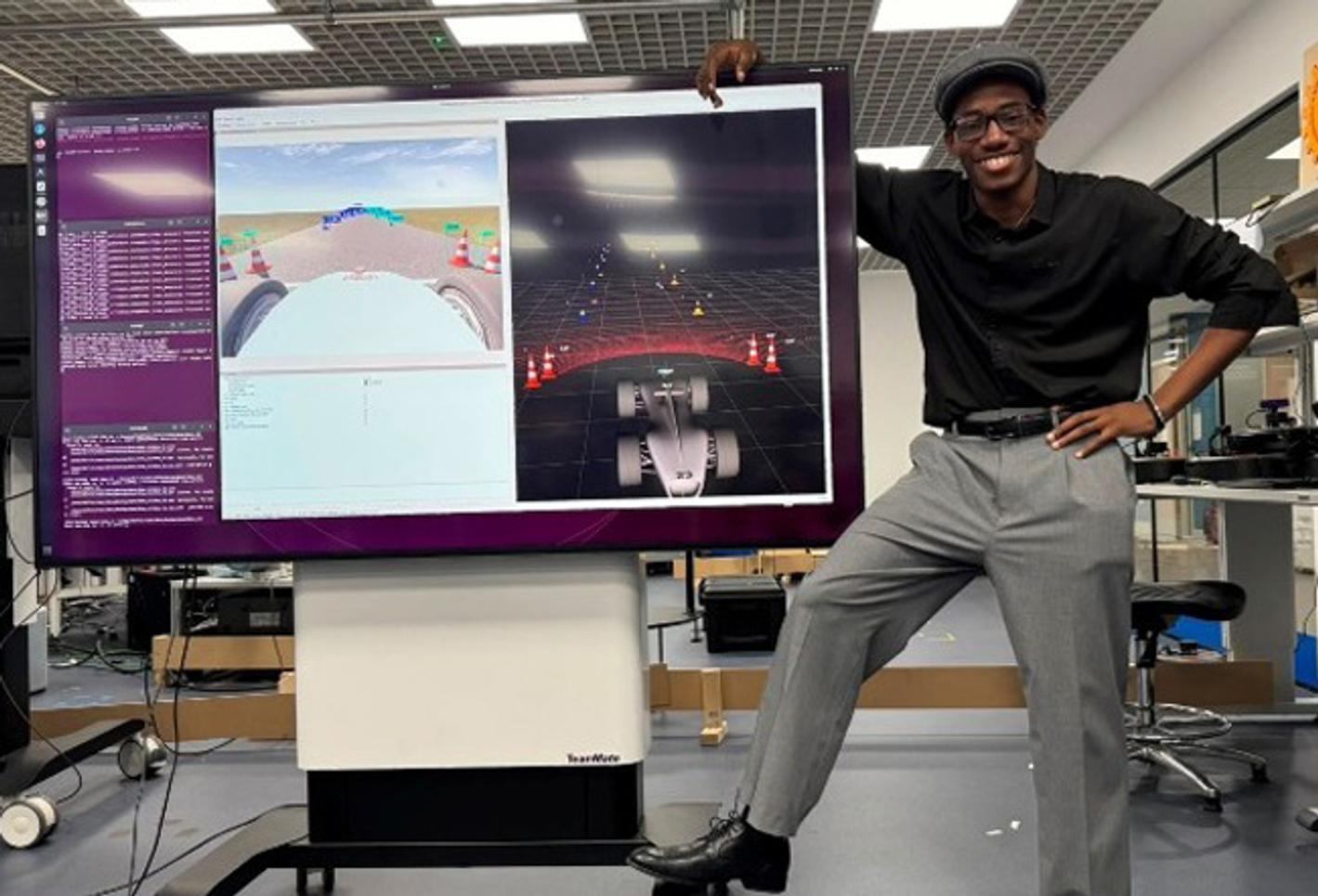 A man in a cap and glasses stands leaning against a large computer monitor, looking at the camera