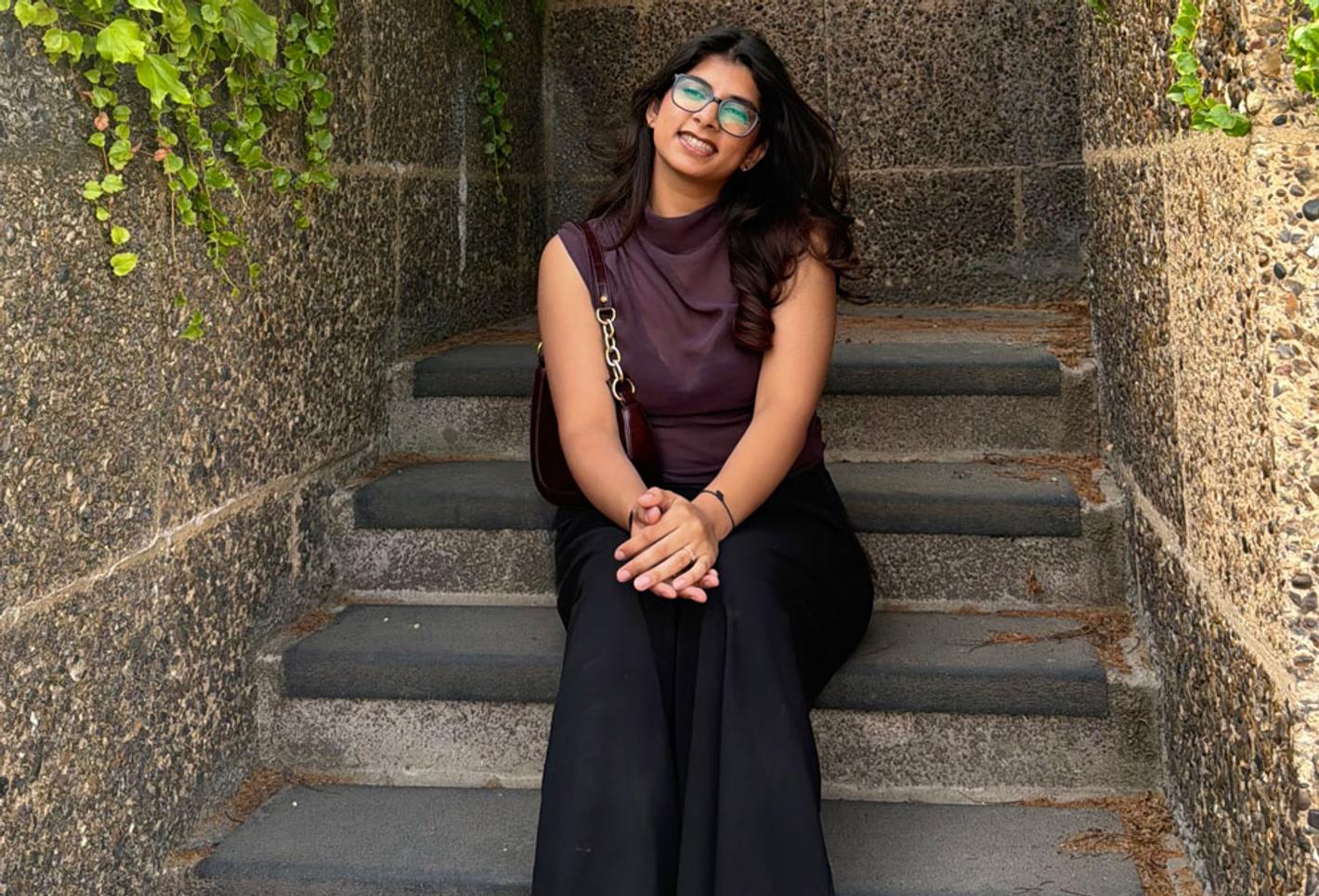 Nitika Kapoor, BA (Hons) Fashion Communication and Styling student, sitting outside in front of steps, with shrubs in the background.