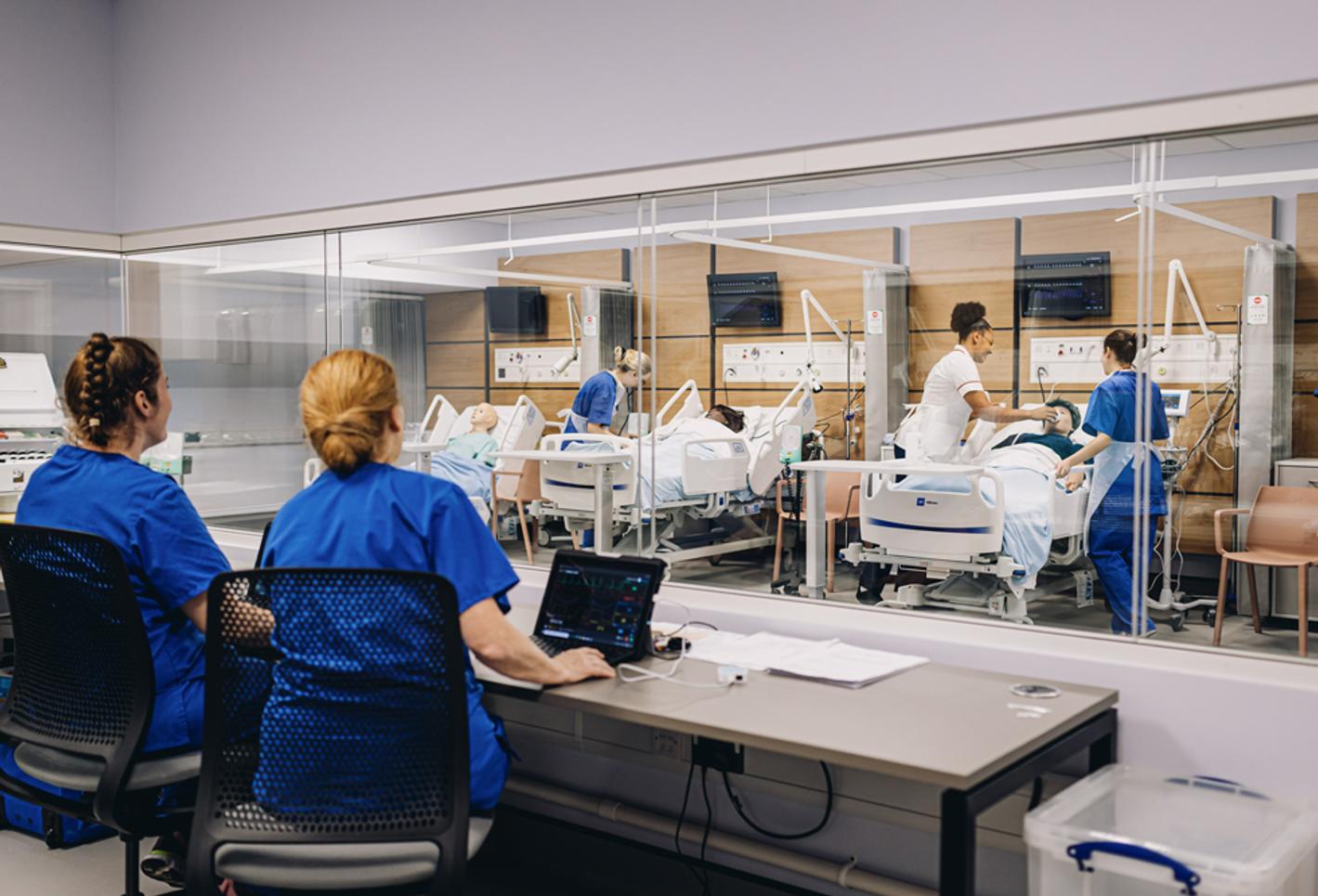a nursing station in a hospital ward with two seated nurses observing the care of patients