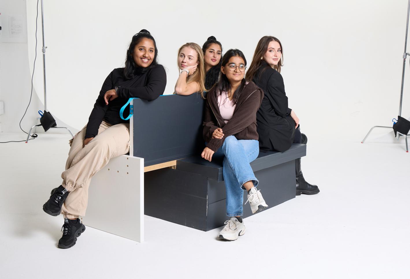 A group of five women sitting on a bench against a white photographic studio backdrop