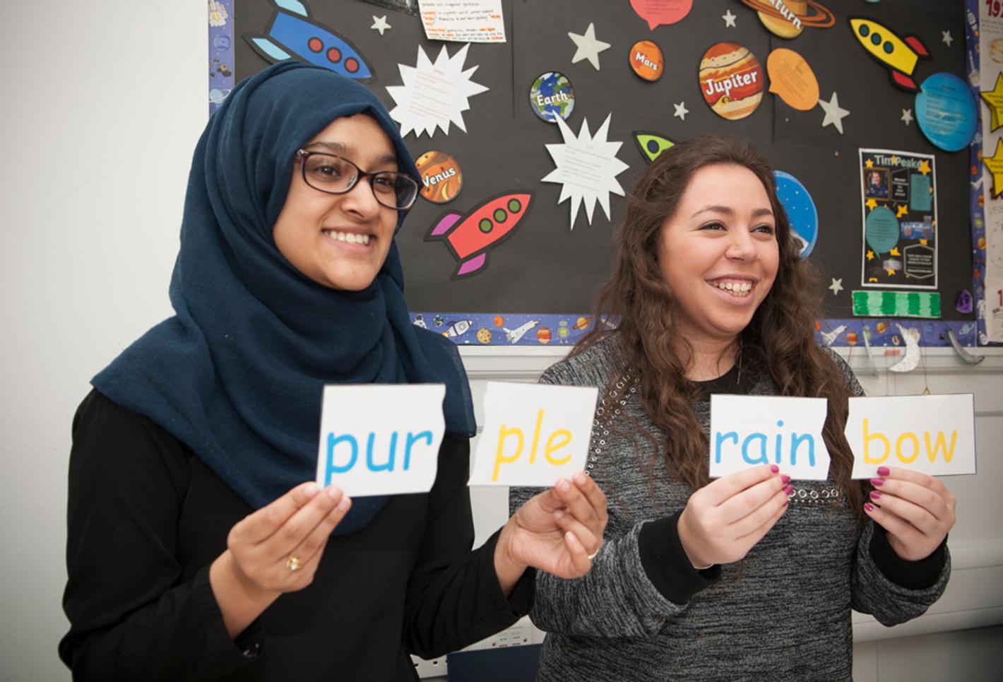 Two women smiling in front of school artwork holding up words on cards