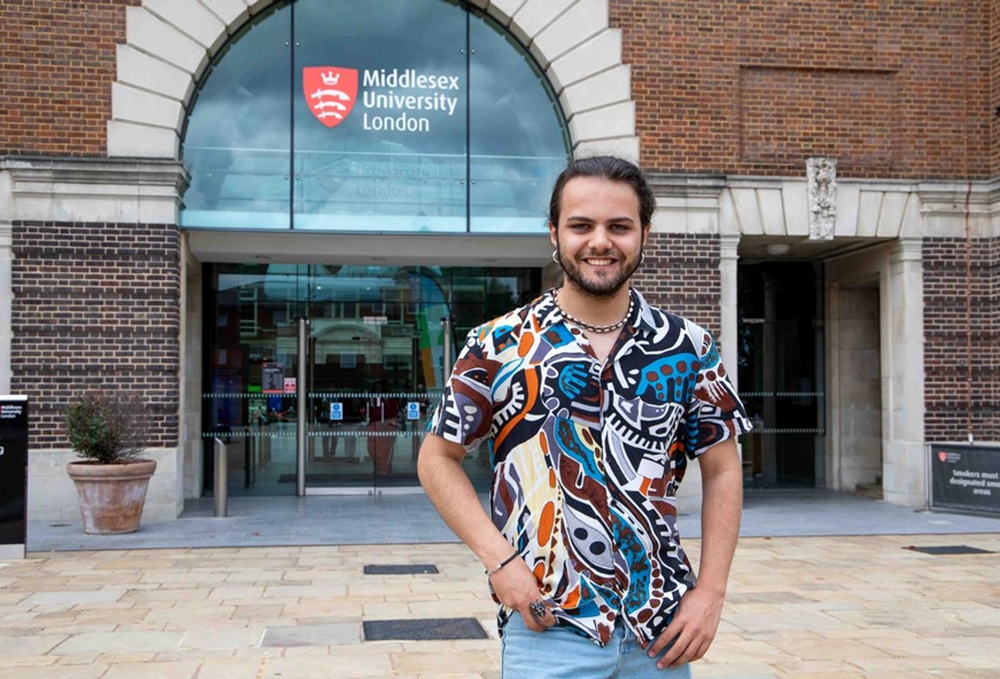 Omar Maaouan Veiga in a colourful shirt, standing and smiling outside the entrance to Middlesex University's College Building