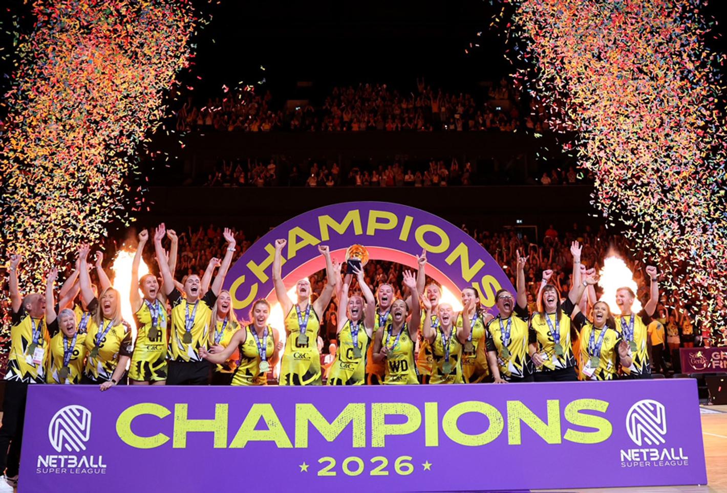 A netball team celebrates behind a sign saying 'champions' and a firework explosion a each side