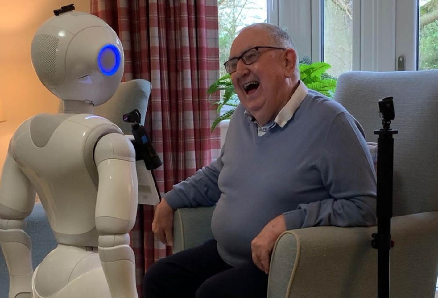 An elderly man sitting in a chair interacting with a robot.
