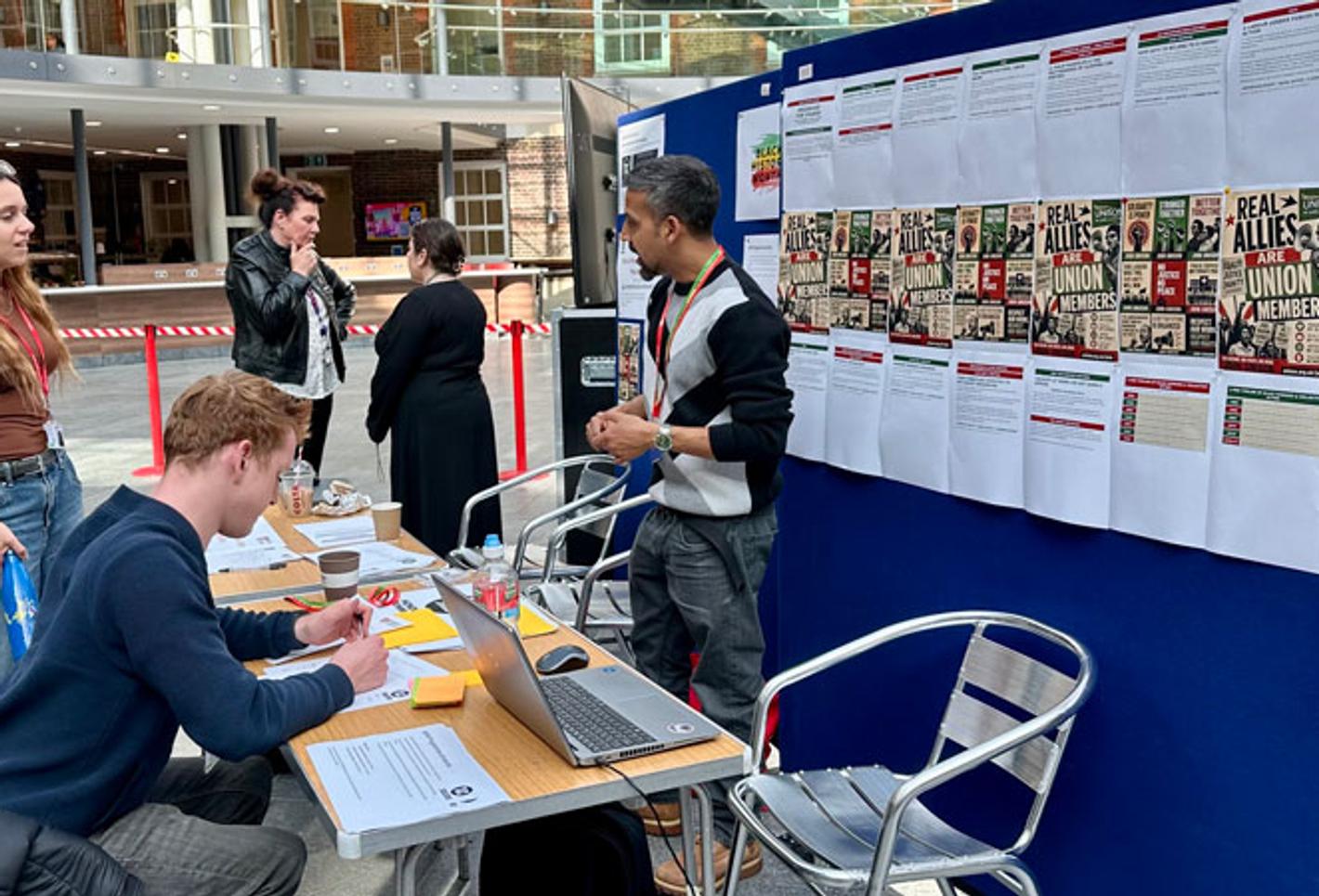 Students at a Black History Month stall, with posters displayed on a board, students seated at a desk and others chatting in the background.