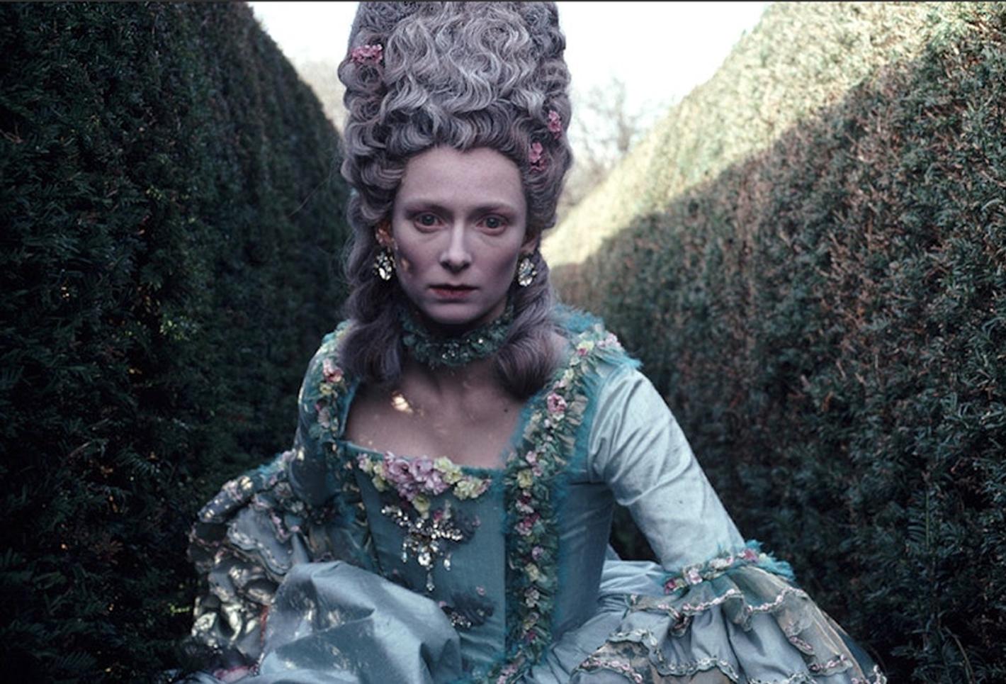 Lady wearing a big hair like Marie Antoinette running through a hedge maze.