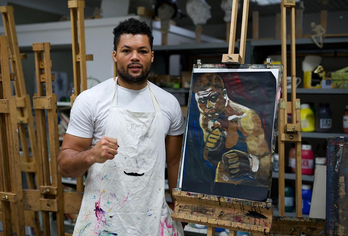 An apron-clad male student displaying a painting of a boxer.
