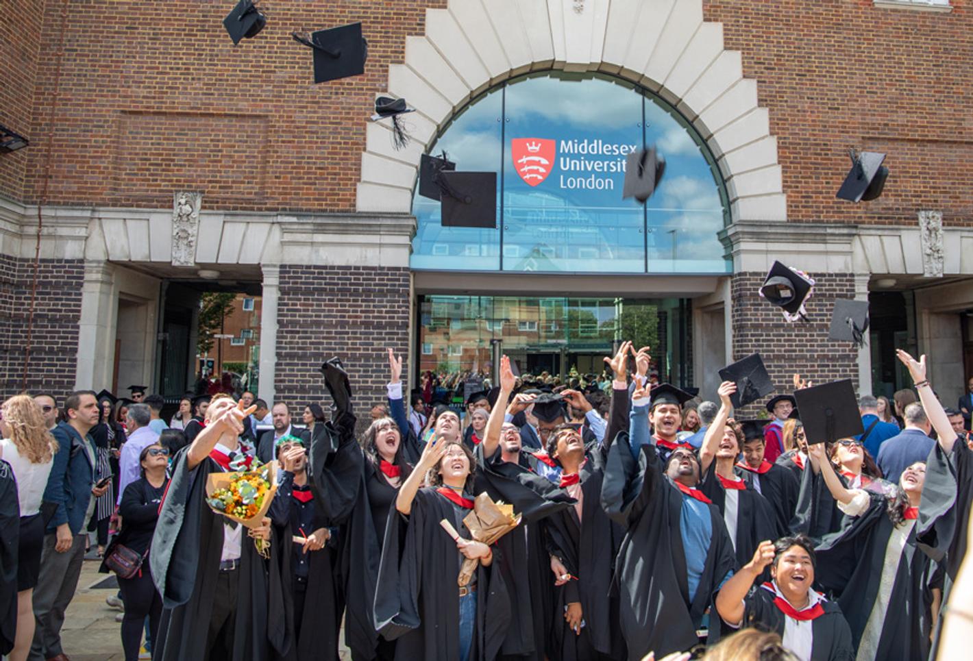 A group of graduates in gowns throw their mortar boards into the air to celebrate graduation in front of a Middlesex University building