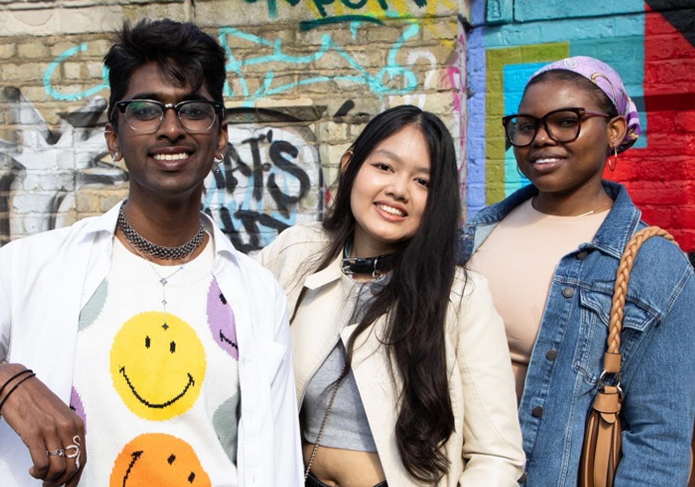 Three smiling students in front of a wall with artistic graffiti in Camden