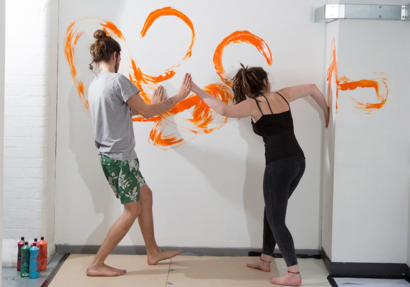 An image of two artists painting with their hands with orange paint on a white wall